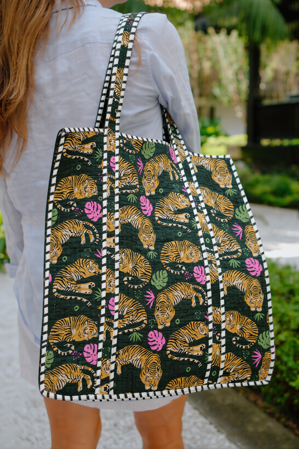 The bag has a dark green base with a repeating pattern of golden-brown leaves and pink flowers, bordered by white and black pom-pom trim.